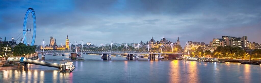 Panoramic night view of London along the Thames with the illuminated London Eye on the left and Westminster landmarks across the water.